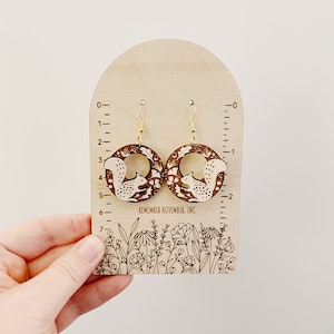 May include: A pair of wooden earrings with a squirrel design. The earrings are brown with white accents and are hanging from gold hooks. The earrings are displayed on a wooden stand with a ruler and the text "REMEMBER NOVEMBER, INC."