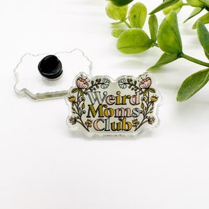 May include: A clear acrylic pin with the text "Weird Moms Club" in a floral design. The pin has a black backing and a pin clasp. The design includes pink, yellow, and green colors. The pin is rectangular with rounded edges.