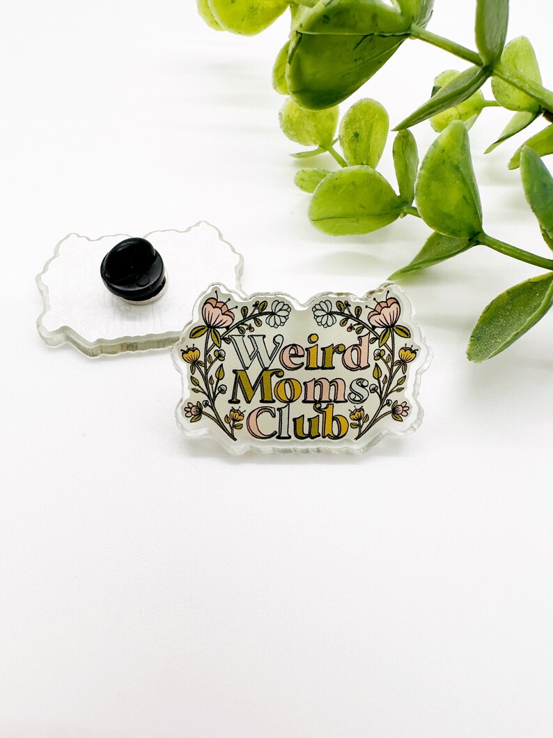 May include: A clear acrylic pin with the text "Weird Moms Club" in a floral design. The pin has a black backing and a pin clasp. The design includes pink, yellow, and green colors. The pin is rectangular with rounded edges.