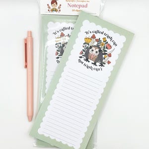 May include: A notepad with a light green background and a scalloped edge, featuring a cartoon opossum illustration with the text "It's called trash can, not trash can't." A pink pen is next to the notepad.