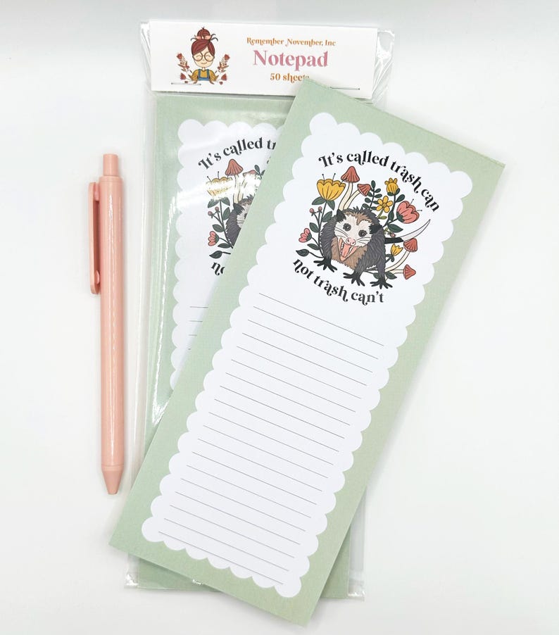 May include: A notepad with a light green background and a scalloped edge, featuring a cartoon opossum illustration with the text "It's called trash can, not trash can't." A pink pen is next to the notepad.