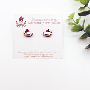 May include: A pair of white and orange miniature cake earrings with red and black accents. The earrings are handmade with love by Remember November, Inc. and are displayed on a white card with the company's website address.