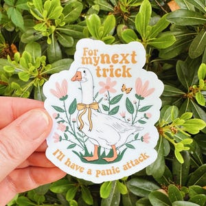 May include: A white sticker featuring a cartoon goose with a yellow bow, surrounded by pink flowers and green foliage. The sticker has the text "For my next trick I'll have a panic attack" in orange and black.