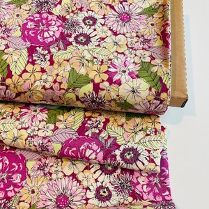 May include: Pink fabric with a floral print featuring yellow, pink, and green flowers. The fabric is folded and stacked on a brown cardboard box.