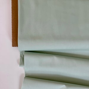 May include: A roll of light green fabric folded on a brown surface. The fabric has a smooth, slightly textured surface.