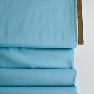 May include: A bolt of solid light blue fabric wrapped around a brown cardboard core. The textile features a smooth, matte finish and is folded in horizontal layers, displaying a consistent, vibrant sky blue color across the entire surface.