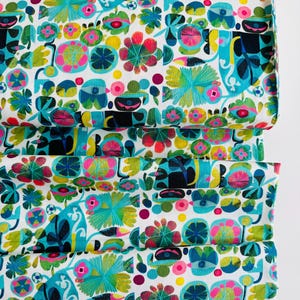 May include: A folded fabric with a vibrant floral pattern. The design features a white background with colorful flowers in shades of teal, pink, yellow, and black. The fabric is likely cotton or a similar material, suitable for sewing projects.