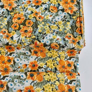 May include: A folded fabric with a vibrant floral pattern. The design features large orange flowers with dark centers, interspersed with smaller yellow and white blossoms on a light green and white background. The fabric is folded, showing the pattern's repeat.