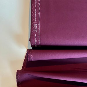 May include: A folded piece of deep burgundy fabric with a white label that reads "#WE ARE Fabrics | galleryfabrics.com | 100% PREMIUM COTTON | STANDARD 100 | OEKO-TEX® | 180001 Certified".
