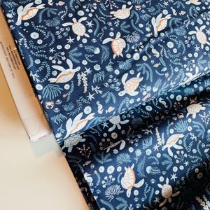 May include: Navy blue fabric with a repeating pattern of sea turtles, seahorses, and aquatic plants in white, light blue, and peach. The fabric is folded, showing the detailed design and texture.