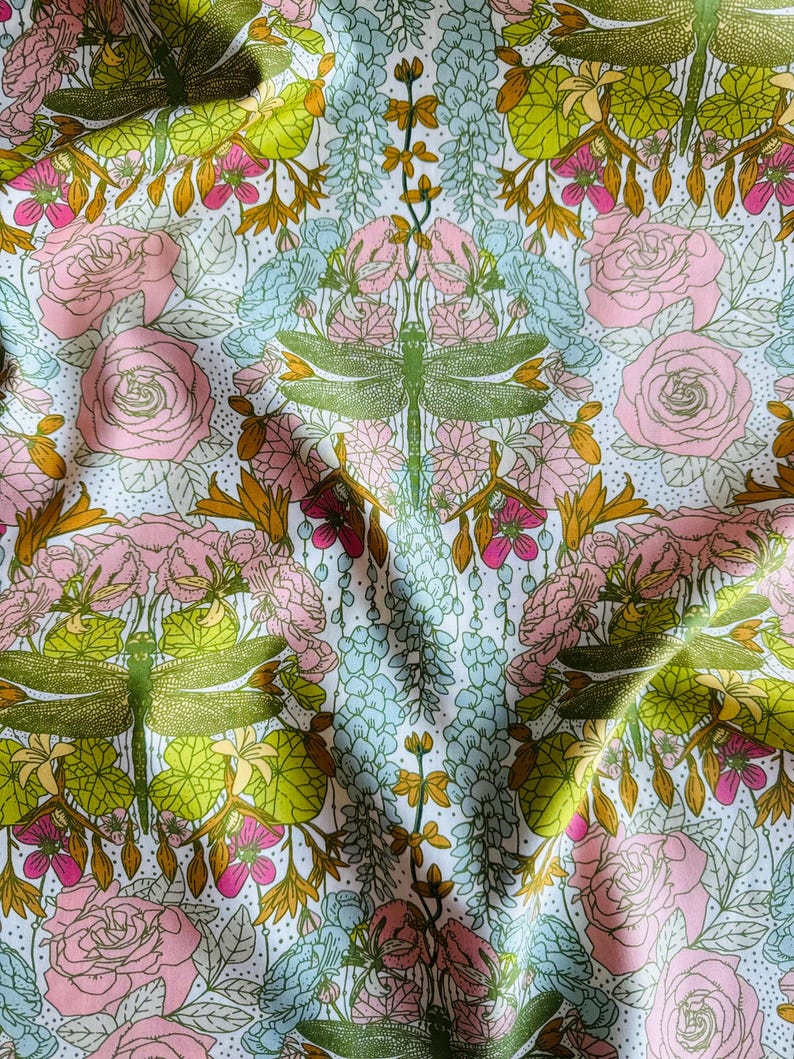 Peut inclure: Un tissu pr&eacute;sentant un motif r&eacute;p&eacute;titif de libellules vertes, de roses roses et de grappes florales bleu clair sur un fond blanc avec de petits points noirs. Le design inclut des feuilles et des tiges vert-jaune dispos&eacute;es sym&eacute;triquement.