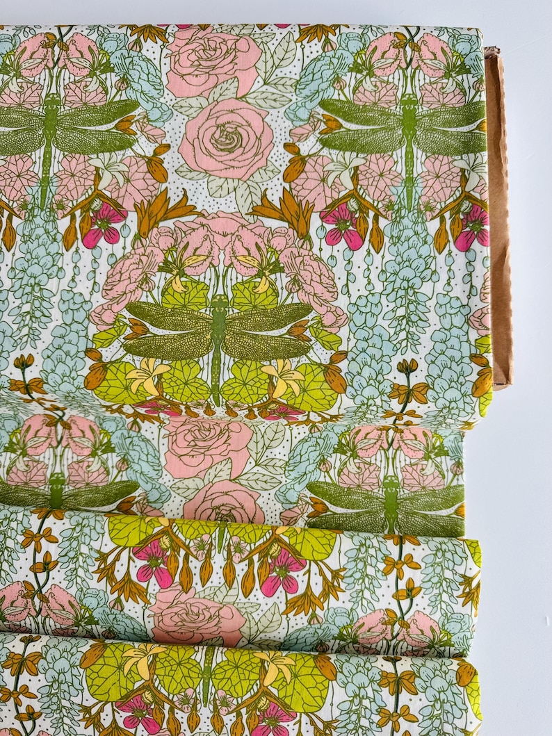 Peut inclure: Un rouleau de tissu pr&eacute;sentant un motif r&eacute;p&eacute;titif de libellules vertes, de roses roses et de grappes de fleurs bleu clair sur un fond blanc. Le design comprend des lignes complexes et des &eacute;l&eacute;ments botaniques vibrants dispos&eacute;s sym&eacute;triquement.