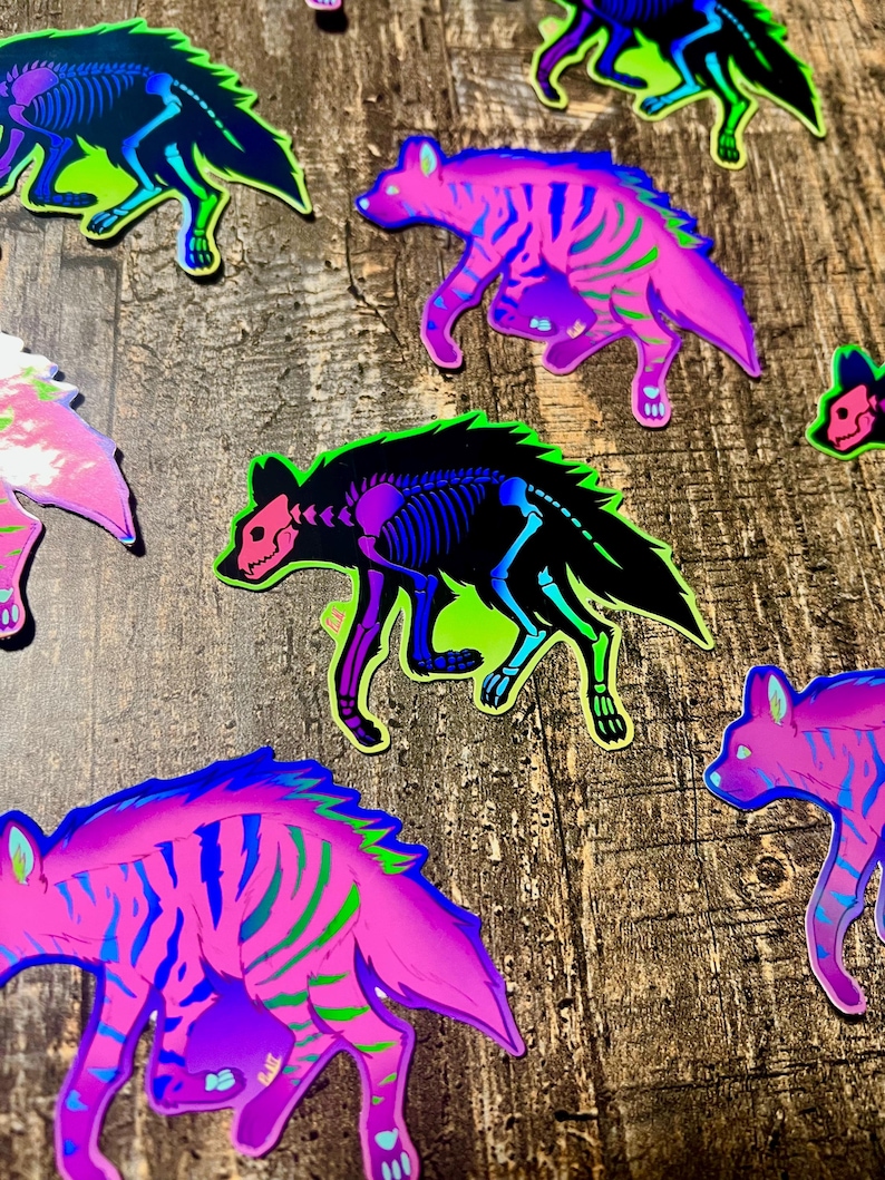 May include: A collection of colorful hyena stickers with a unique design. The stickers feature a hyena silhouette with a skeleton overlay, in shades of pink, blue, green, and black. The stickers are on a wooden surface.