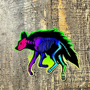 May include: A holographic sticker featuring a stylized hyena with a skeleton design. The hyena's skull is pink, the rib cage is purple and blue, and the outline is neon green. The sticker is on a wooden surface.