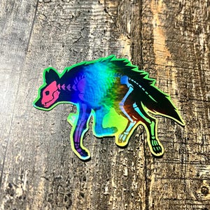 May include: Holographic sticker featuring a stylized hyena skeleton. The sticker has a black outline with a rainbow iridescent fill. The hyena's skull and skeletal structure are pink and white. The sticker is on a wooden surface.