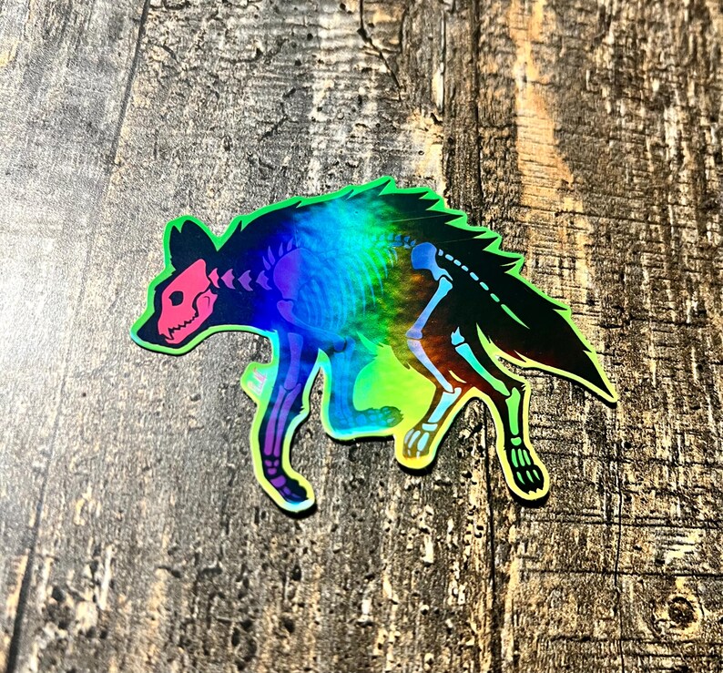 May include: Holographic sticker featuring a stylized hyena skeleton. The sticker has a black outline with a rainbow iridescent fill. The hyena's skull and skeletal structure are pink and white. The sticker is on a wooden surface.