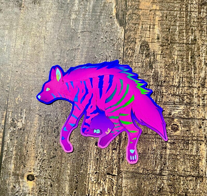 May include: A vibrant sticker featuring a stylized hyena in shades of pink, blue, and green. The hyena has striped markings and is depicted in a walking pose. The sticker is on a wooden surface.