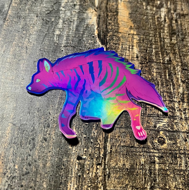 May include: A holographic sticker featuring a stylized hyena in shades of purple, blue, and green. The sticker has a rainbow sheen and is depicted in a walking pose. The sticker is on a wooden surface.
