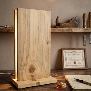 Może przedstawiać: Drewniana lampa stołowa o prostokątnym kształcie. Lampa ma jasne drewniane wykończenie i pionowy pasek świetlny z boku. Lampa stoi na drewnianej podstawie. Drewniana półka z narzędziami i oprawiony certyfikat znajdują się w tle.