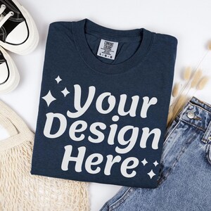 May include: Navy blue t-shirt with the text "Your Design Here" in white. The shirt is folded and has a Comfort Colors tag. A pair of sneakers and denim jeans are also in the image.