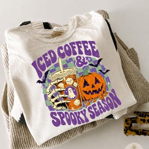 May include: A cream-colored t-shirt featuring a graphic of a skeleton hand holding an iced coffee cup next to a jack-o'-lantern. The design includes a purple and green checkered background, small bats, and the text ICED COFFEE & SPOOKY SEASON.