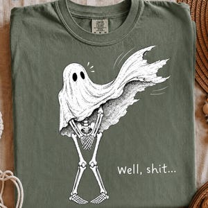 May include: An olive green t-shirt featuring a white graphic illustration of a skeleton wearing a ghost sheet. The ghost sheet is blowing to the right, revealing the skeleton underneath. The text "Well, shit..." is printed in white below the graphic.