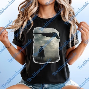 Könnte beinhalten: Schwarzes T-Shirt mit einer Grafik einer Figur in einem Umhang mit einer Waffe, die in einer nebligen Landschaft unter einem dunklen Himmel steht. Eine Raumschiff-Silhouette befindet sich darüber. Das T-Shirt hat kurze Ärmel.