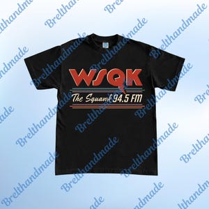 Może przedstawiać: Czarny t-shirt z napisem "WSQK The Squawk 94.5 FM" w kolorze czerwonym, niebieskim i pomarańczowym. Projekt ma styl retro z poziomymi liniami i błyskawicą.