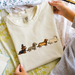 Könnte beinhalten: Ein cremefarbenes T-Shirt mit einer Grafik von vier Dackeln in Halloween-Kostümen. Die Hunde sind als Hexe, Mumie, Hotdog und Geist verkleidet. Das T-Shirt hat ein Markenetikett von Comfort Colors am inneren Halsausschnitt.