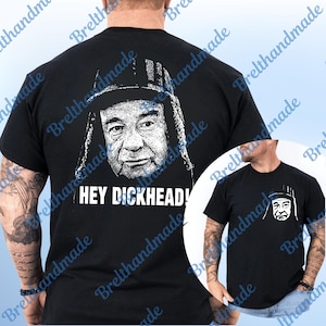Può includere: T-shirt nero con grafica bianca di un uomo con cappello e la scritta "HEY DICKHEAD!" in bianco. Il disegno è sul davanti e sul retro. La t-shirt è a maniche corte.