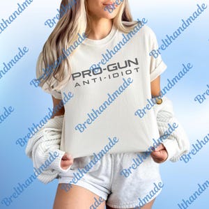 May include: A cream-coloured t-shirt featuring the text "PRO-GUN ANTI-IDIOT" in dark grey. The shirt has rolled-up sleeves and a relaxed fit. The design is straightforward and bold.