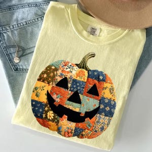 Könnte beinhalten: Ein hellgelbes Kurzarm-T-Shirt mit einer großen Grafik eines Halloween-Kürbisses, der aus einem Patchwork-Muster mit verschiedenen Blumen- und Strukturmustern in Orange, Blau und Grün besteht, sowie einem schwarzen Gesicht.