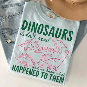 May include: A light blue t-shirt featuring dark green text that reads "DINOSAURS didn't read and we know what HAPPENED TO THEM" along with pink line-art illustrations of four different dinosaurs arranged in the center of the garment.