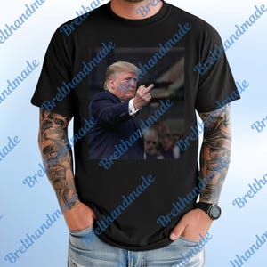 May include: Black t-shirt featuring a square image of a man in a suit extending his middle finger. The image is centered on the shirt. The shirt is worn by a person with tattoos on their arm.