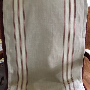 May include: A beige linen fabric with red and white stripes. The fabric is folded and draped over a chair.