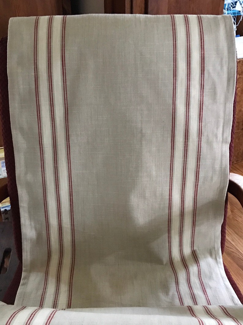 May include: A beige linen fabric with red and white stripes. The fabric is folded and draped over a chair.