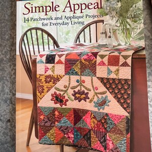 May include: A book titled "Simple Appeal" by Kim Diehl, featuring 14 patchwork and appliqué projects for everyday living. The book cover showcases a colorful quilt with floral motifs and geometric patterns, displayed on a table with a vase of flowers.