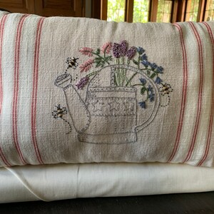 May include: A beige pillow with red stripes features an embroidered design of a watering can filled with flowers and bees.
