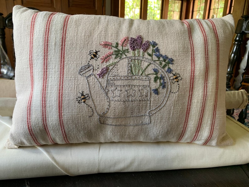May include: A beige pillow with red stripes features an embroidered design of a watering can filled with flowers and bees.