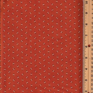 May include: Rust-colored fabric with a repeating pattern of small white and black floral designs. A wooden ruler with inch measurements is visible along the right edge of the fabric. The fabric is likely cotton or a similar material.