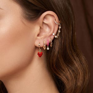 May include: A collection of gold-toned earrings worn in a single ear, featuring a small hoop with a red strawberry charm, a textured gold hoop, a solid pink hoop, a small chain link hoop, and a gold ear cuff connected by a delicate chain.