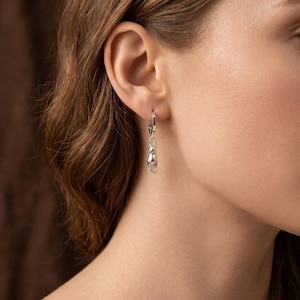 May include: Silver cat-shaped dangle earring. The earring features a small, detailed cat pendant suspended from a hoop. The cat is depicted in a stylized, minimalist design. The earring is shown on a person's ear, highlighting its size and design.