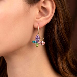 May include: A gold-toned dangle earring featuring a butterfly-shaped design composed of marquise-cut gemstones in purple, blue, green, and pink. The earring includes a small hook with embedded clear stones and two thin gold wires as antennae.