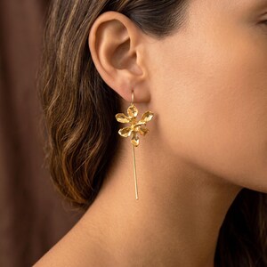 May include: A close-up of a gold-colored earring featuring a flower design with six petals. The earring hangs from a thin, straight gold bar. The earring is worn on a person's ear.