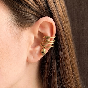 May include: A gold-toned ear cuff shaped like a safety pin with a small dangling skull charm. The jewelry piece wraps around the outer edge of the ear cartilage, featuring a metallic finish and a detailed miniature skull hanging from the bottom loop.