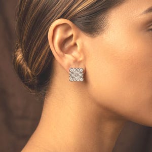 May include: Square silver stud earring with a repeating, openwork design. The earring is adorned with small, sparkling gemstones, creating a delicate and elegant appearance. The earring is shown on a person's ear, highlighting its size and style.