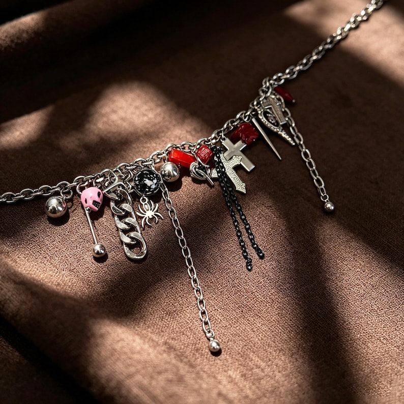 K&ouml;nnte beinhalten: Eine silberne Halskette mit verschiedenen Anh&auml;ngern, darunter ein rosa Sch&auml;del, eine Spinne, ein Kreuz und rote Perlen. Die Halskette hat eine dunkle, gotische &Auml;sthetik mit einer Mischung aus Texturen und Formen.