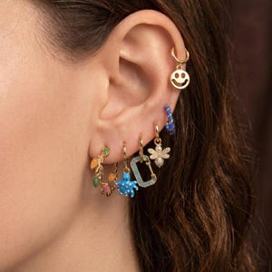 May include: A set of six gold-tone hoop earrings featuring various charms including a smiley face, a blue spiral hoop, a bee with white stones, a rectangular link, a blue octopus, and a hoop with multi-colored teardrop-shaped stones.