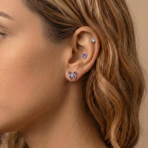 May include: A close-up of a person's ear adorned with three earrings. The lower lobe features a butterfly stud earring with purple and white stones. Above, a small purple stud and a clear crystal stud are visible.