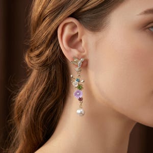 Può includere: Un orecchino pendente color oro con un colibrì nella parte superiore, seguito da una disposizione verticale di piccoli ciondoli floreali multicolori in blu, verde, viola e rosa, che termina con una perla sferica bianca alla base.